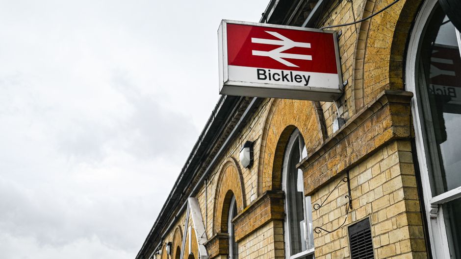 A close-up view of a brick railway station building featuring arched windows, with a prominent red and white 'Bickley' sign hanging from the exterior. The sign has the iconic British railway symbol an