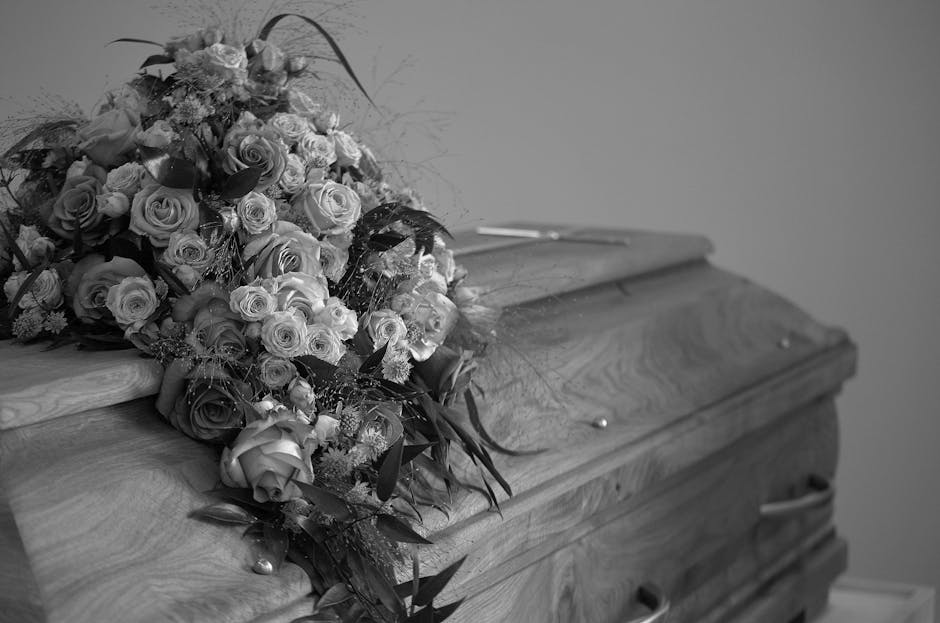 A large, meticulously arranged funeral floral tribute placed atop a wooden casket. The bouquet features a combination of roses in various shades, with some fully bloomed and others in bud, complemente
