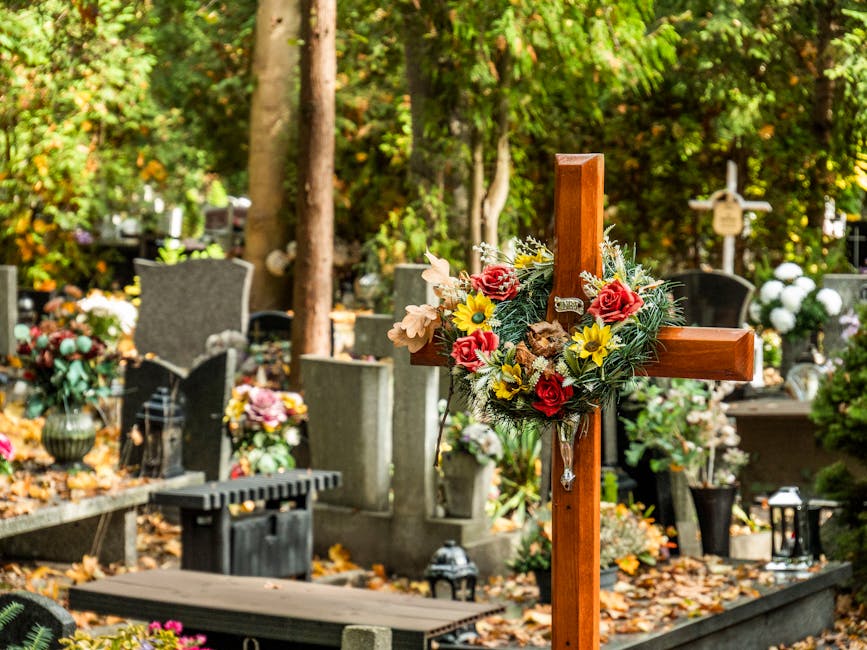 A wooden cross adorned with a vibrant floral arrangement featuring pink roses, yellow daisies, white lilies, and greenery, situated in a cemetery surrounded by gravestones, trees, and fallen autumn le