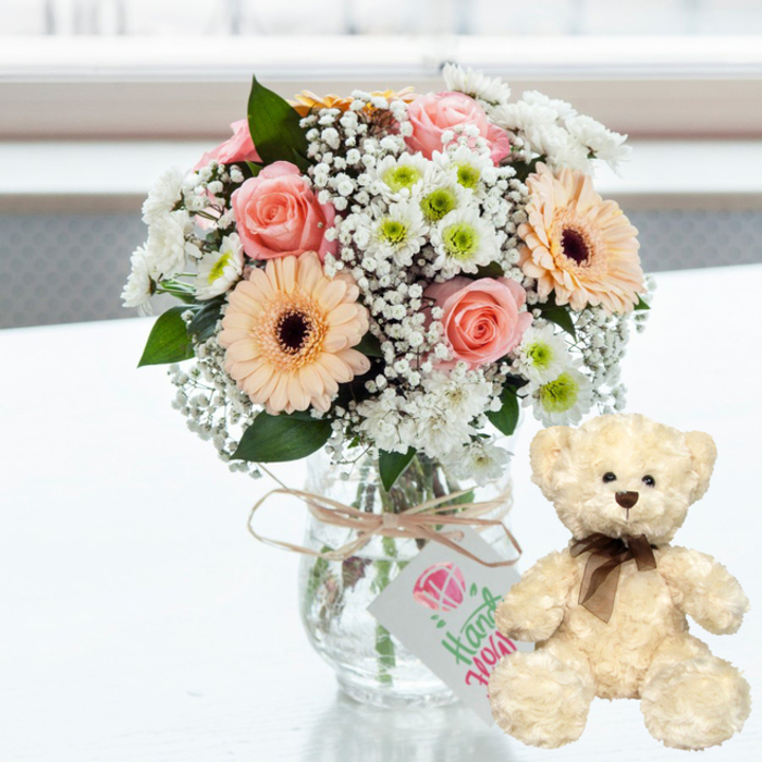 A soft, romantic still life is set against a bright, softly blurred background, capturing the feeling of an intimate anniversary gesture in Norbiton. In the centre, a rounded bouquet of pastel flowers nestles in a clear glass jar-style vase, its transparency allowing the stems and water to be seen clearly. The arrangement brings together blush pink roses with velvety petals, peach-toned gerbera daisies featuring deep chocolate-brown centres, and airy sprays of white baby's breath that thread between the larger blooms. Small white chrysanthemums with lively lime-green centres add a crisp freshness, while scattered hints of dark green foliage give the design balance and depth, much like the mixed planting you might see in a well-tended garden off Gloucester Road. Around the vase's neck, a length of natural twine is tied into a modest bow, supporting a dangling white rectangular tag. On the tag, a pink heart emblem sits above a graceful line of green script that reads "Handmade," gently reinforcing the sense of personal care and local craftsmanship behind the gift. To the right of this floral focal point, an endearing cream-coloured teddy bear sits upright, angled slightly towards the viewer. Its fur appears soft and textured, inviting touch, while its dark button eyes, brown stitched nose, and slim brown satin ribbon at the neck add a timeless, comforting character. The smooth white tabletop reflects a faint echo of both vase and bear, enhancing the scene's lightness and clarity. Bathed in natural daylight that suggests a nearby window, this pairing of flowers and teddy bear feels perfect for commemorating an anniversary, sending love from Norbiton to someone special, or creating a cosy, sentimental corner in a shared home.